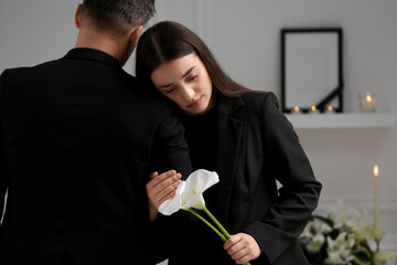 Sad couple with calla lily flowers mourning indoors, space for text. Funeral ceremony