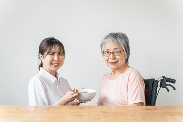 老人ホーム・家で食事する笑顔の高齢者女性と食事介助する管理栄養士・介護士・ヘルパーのアジア人女性
