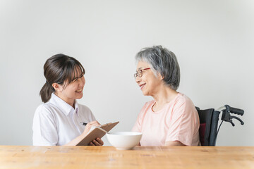 老人ホーム・家で食事する笑顔の高齢者女性と食事介助する管理栄養士・介護士・ヘルパーのアジア人女性
