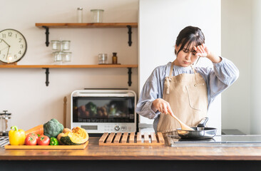 料理中に体調不良になるエプロン姿の女性・主婦（頭痛）
