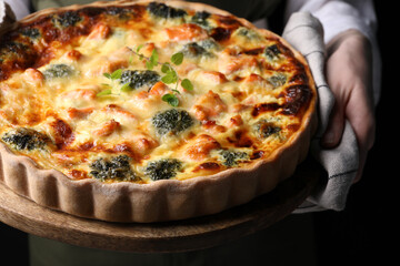 Woman holding wooden board with delicious homemade salmon quiche on black background, closeup