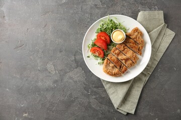 Tasty cut schnitzel served with sauce, microgreens and tomato on grey textured table, top view....