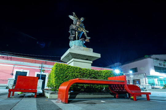 Tegucigalpa, Francisco Morazan, Honduras - December 11, 2022: Statue of Saint Michael Archangel at Night in "Los Dolores" Square on a Pedestal with Red Benches Around