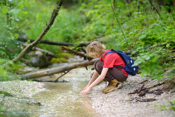 Preteen boy in red shirt is exploring nature and playing with water in brook during hiking in mountains valley. Active leisure for energetic children. Summer outdoor recreation for kids - obrazy, fototapety, plakaty