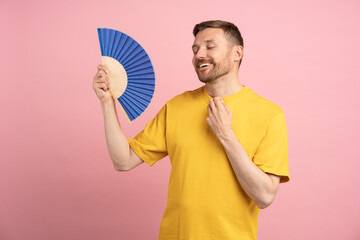 Carefree smiling man using paper fan at summer hot weather, feel relief from heat, cools herself,...