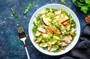 Waldorf salad with fresh apple, celery, lettuce, grilled chicken fillet, arugula and walnuts on plate, blue table background, top view