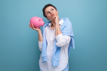 a pensive young woman with a short haircut holds a piggy bank and thinks where to invest the...