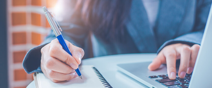 Woman hand is writing on a notepad with a pen and have a laptop computer on the desk in the office.