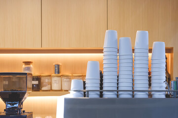 Rows of many white paper cups on coffee maker with grinder in front of wooden shelf of coffee kiosk in food court area 