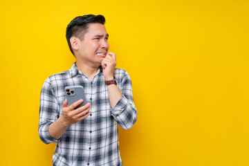 Worried young Asian man wearing a white checkered shirt holding smartphone and biting his nails, looking side with a frustrated expression isolated over yellow background. People Lifestyle Concept