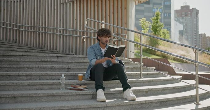 Young indian guy student reading book sitting on a stairs outdoor