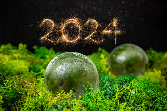 2024 number written with golden sparklers next to glass spheres on green lichen with black background, - Powered by Adobe