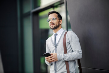 A young smart casual businessman is standing in front of the business center and holding his phone.