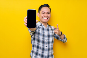 Smiling handsome young Asian man wearing a white checkered shirt pointing finger at mobile phone with blank screen, Advertising new mobile app isolated over yellow background. People Lifestyle Concept
