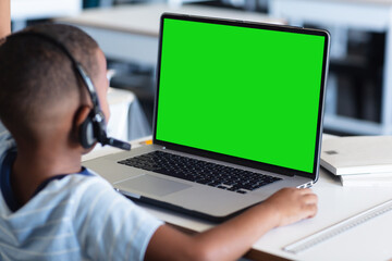 African american boy wearing headphones and studying online over laptop on table at home