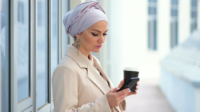 Muslim woman in silver hijab drinks coffee scrolling social media on smartphone. Businesswoman enjoys looking through news update outside office