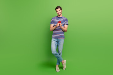 Full length photo of positive cute guy wear striped t-shirt communicating modern gadget isolated green color background