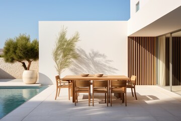 On a sunny day, a valuable modern minimalist villa is adorned with a dining table, chairs, and wooden sun loungers near a swimming pool in its backyard.
