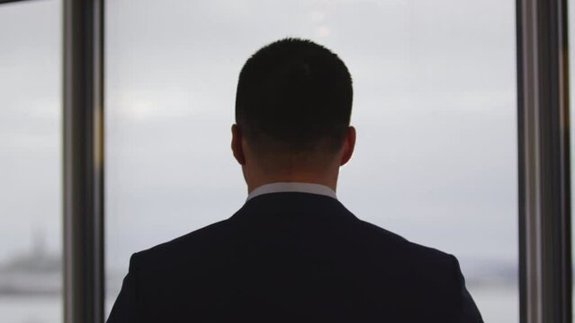 Young man stands in official dark suit and white shirt near office window at noon back view. Male brunet hair cut shortly