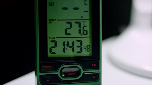 Hand of person turns on digital clock standing at desk in study room at nighttime. Display of clock shows evening time and temperature of room at dark closeup