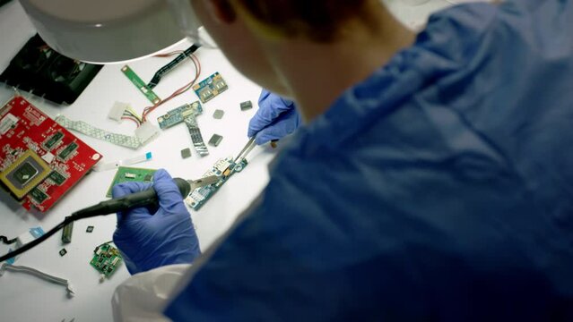 Technical worker in special clothes and gloves solders motherboard of electronic device under led lamp. Lamp light exposes details and wire parts of mainboard