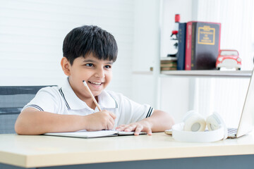 Smiling Indian handsome child boy writing taking notes in note book for studying, near laptop and headphones at home. Online learning. education concept