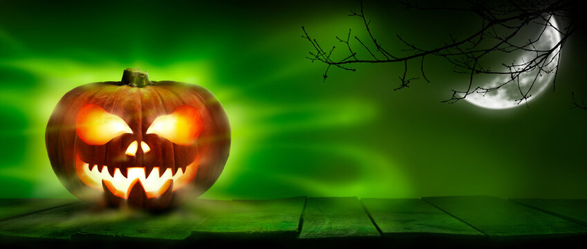 A spooky halloween pumpkin, Jack O Lantern, with an evil spinning green aura glow on a wooden table against a dark background with a crescent moon and tree silhouette.