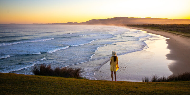 Beautiful girl in yellow dress on green hill admires colorful sunset on the coast. Look At Me Now Headland Walk, Emerald Beach near Coffs Harbour, NSW, Australia	