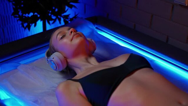 Young pretty woman with headphones lies down in the sensory deprivation chamber, neon light, relax in spa, meditation.