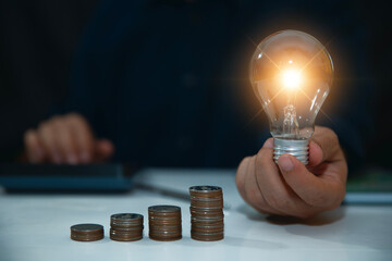 businessman hand holding lightbulb with coins stacking on desk.saving energy and money concept.