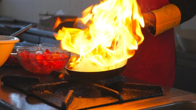 Male chef cooking flambe style dish on pan in modern kitchen of restaurant. Professional cook tossing and frying vegetables in flaming skillet on gas plate. Meal preparation concept. Slow motion
