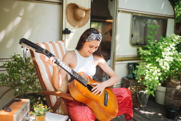 Happy hippie girl is having a good time with playing on guitar in camper trailer. Holiday,...