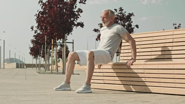 Full shot of senior gray-haired man in sportswear doing triceps dips by street bench outdoors in city during morning workout