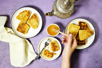 hands eating a harcha or mbasses semolina algerian pancakes on an arabesque colorful plate above cutting board. Traditional algerian breakfast bread with honey bowl