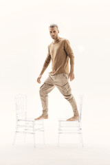 A guy in a beige jumper and cargo pants stands on transparent chairs on a white background.