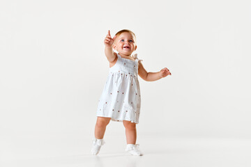 Little smiling baby girl, toddler in beautiful dress walking and raising finger in curiosity over white studio background. Concept of childhood, newborn lifestyle, happiness, care. Copy space for ad