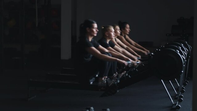 Four Slender Women Doing Exercise On Rowing Machine In Fitness Center, Sport And Healthy Lifestyle