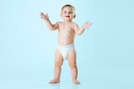 Portrait of little baby girl, toddler in diaper standing and clapping hands against light blue studio background. Cheerful. Concept of childhood, newborn lifestyle, happiness, care. Copy space for ad