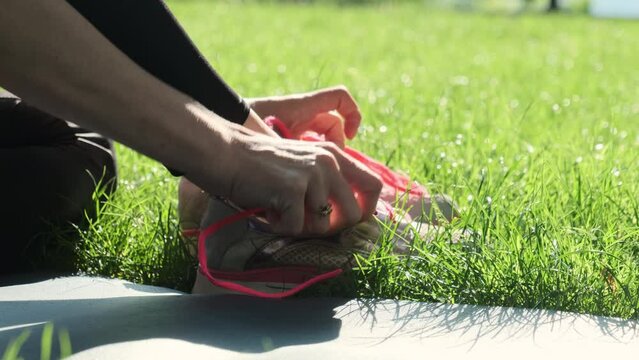 Female person takes shoes with pink laces off during morning yoga practice in city park. Woman unlaces pink trainers sitting on grass slow motion