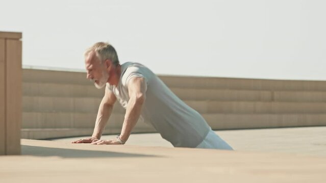Muscular senior man in sportswear practicing push ups at urban public space while working out outdoors in morning
