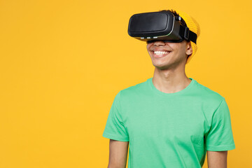 Young smiling fun man of African American ethnicity he wears casual clothes green t-shirt hat watching in vr headset pc gadget isolated on plain yellow background studio portrait. Lifestyle concept.