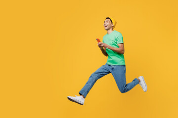 Fototapeta na wymiar Full body side view young man of African American ethnicity he wears casual clothes green t-shirt hat jump high hold in hand use mobile cell phone run fast isolated on plain yellow background studio.