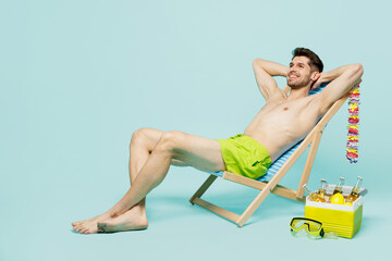 Fototapeta na wymiar Full body side view young sexy man wear green shorts swimsuit relax near hotel pool sit in deckchair hold hands behind neck isolated on plain blue background. Summer vacation sea rest sun tan concept.