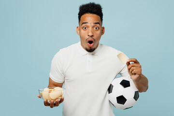 Young shocked surprised amazed man fan wear basic t-shirt cheer up support football sport team hold in hand soccer ball eat chips in bowl watch tv live stream isolated on plain blue color background.