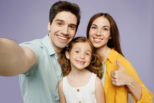 Close up young fun happy parents mom dad with child kid daughter girl 6 year old wear blue yellow casual clothes do selfie shot pov mobile cell phone show thumb up isolated on plain purple background.