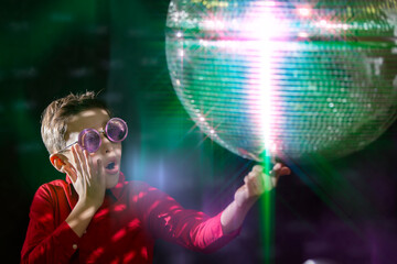 Surprised admiring young boy in stylish glasses smiles with a spinning disco ball.