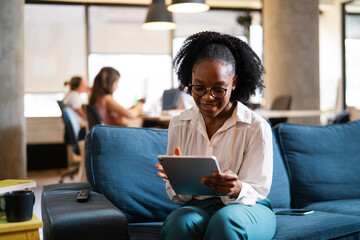 Fototapeta premium Beautiful African businesswoman sitting on sofa in office. Young woman using digital tablet.