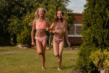 Two little girls with a good tan in a swimsuit in summer play on the field near the garden in the village on a green lawn