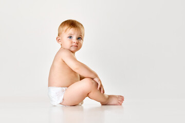Beautiful little baby, toddler in diaper sitting on floor against white studio background. Calm child. Concept of childhood, newborn lifestyle, happiness, care. Copy space for ad - obrazy, fototapety, plakaty