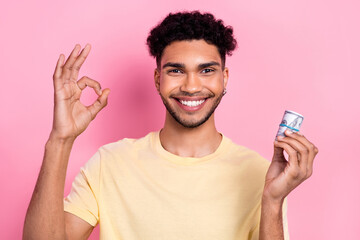 Photo of cheerful toothy beaming guy with earrings dressed yellow t-shirt hold dollars show okey isolated on pink color background